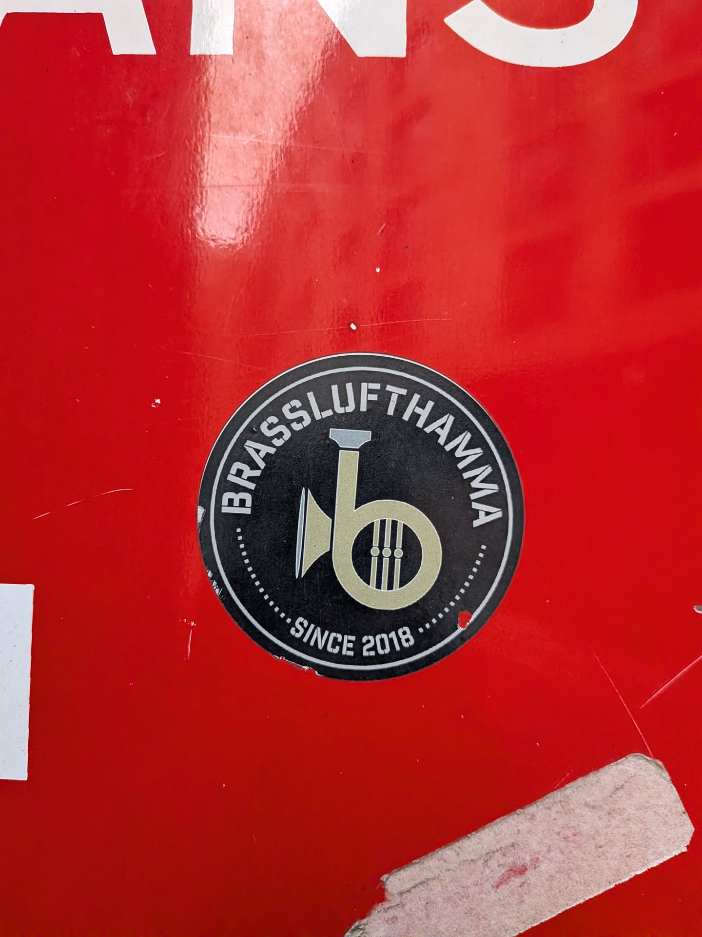 A circular sticker with a black background and a white outer ring. The central design is a stylized, light gold/tan brass instrument, possibly a French horn or tuba bell, with three vertical lines inside its bell. White capital letters spell 'BRASSLUFTHAMMA' along the top curve of the outer ring and 'SINCE 2018' along the bottom curve. A dotted white line separates the black inner circle from the white outer ring. The sticker has a clean, professional, and musical theme.