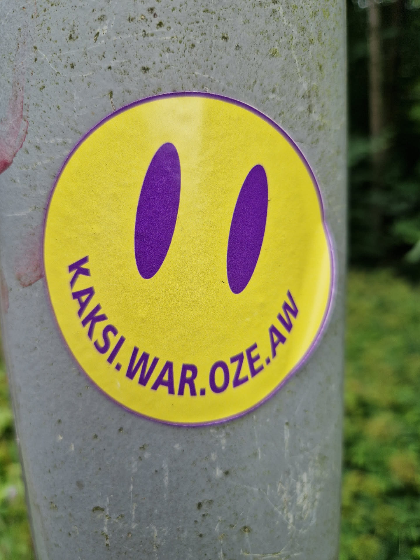 A yellow circle sticker with a purple smiley face and the text "kaksi.war.oze.aw" on it. The sticker is attached to a gray pole.