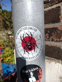 Round sticker featuring a black spider on a red circle surrounded by a spiderweb. The text around the image mentions the artist's Instagram account (@vincentveldhoen and @schiftmacherveldhoen).