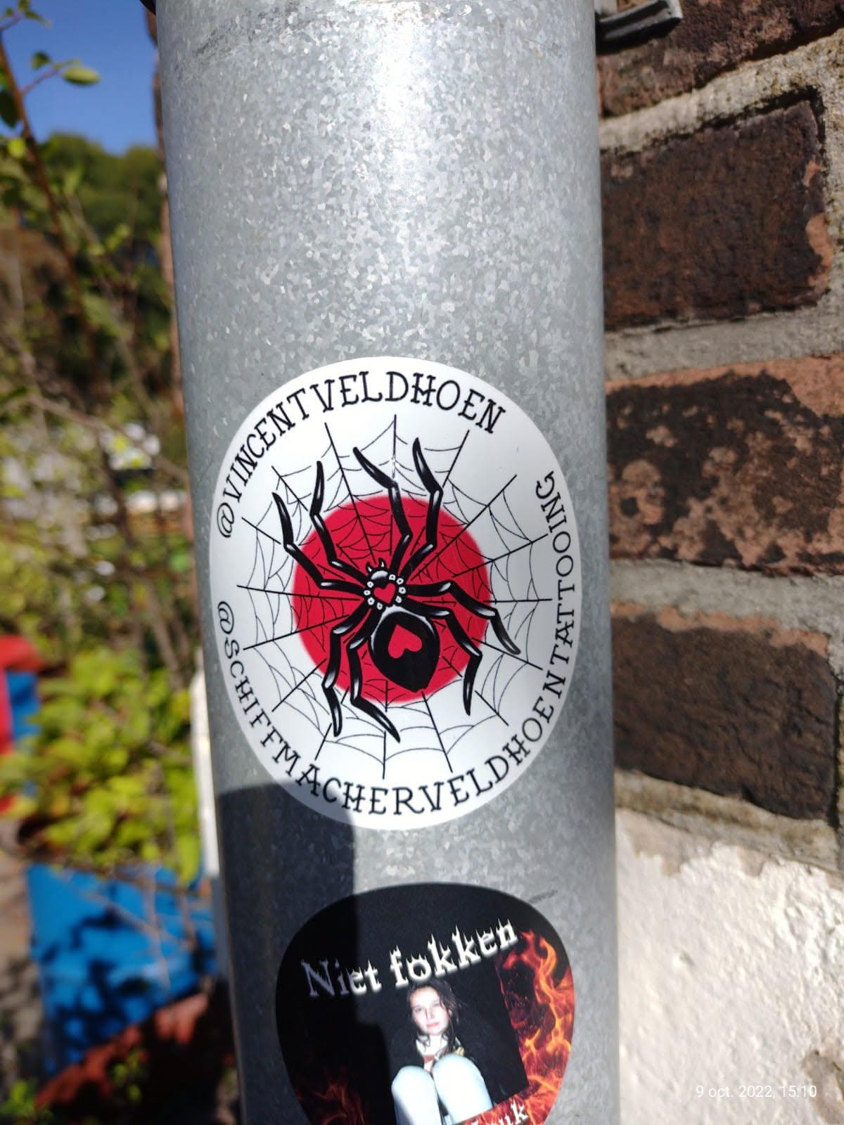 Round sticker featuring a black spider on a red circle surrounded by a spiderweb. The text around the image mentions the artist's Instagram account (@vincentveldhoen and @schiftmacherveldhoen).