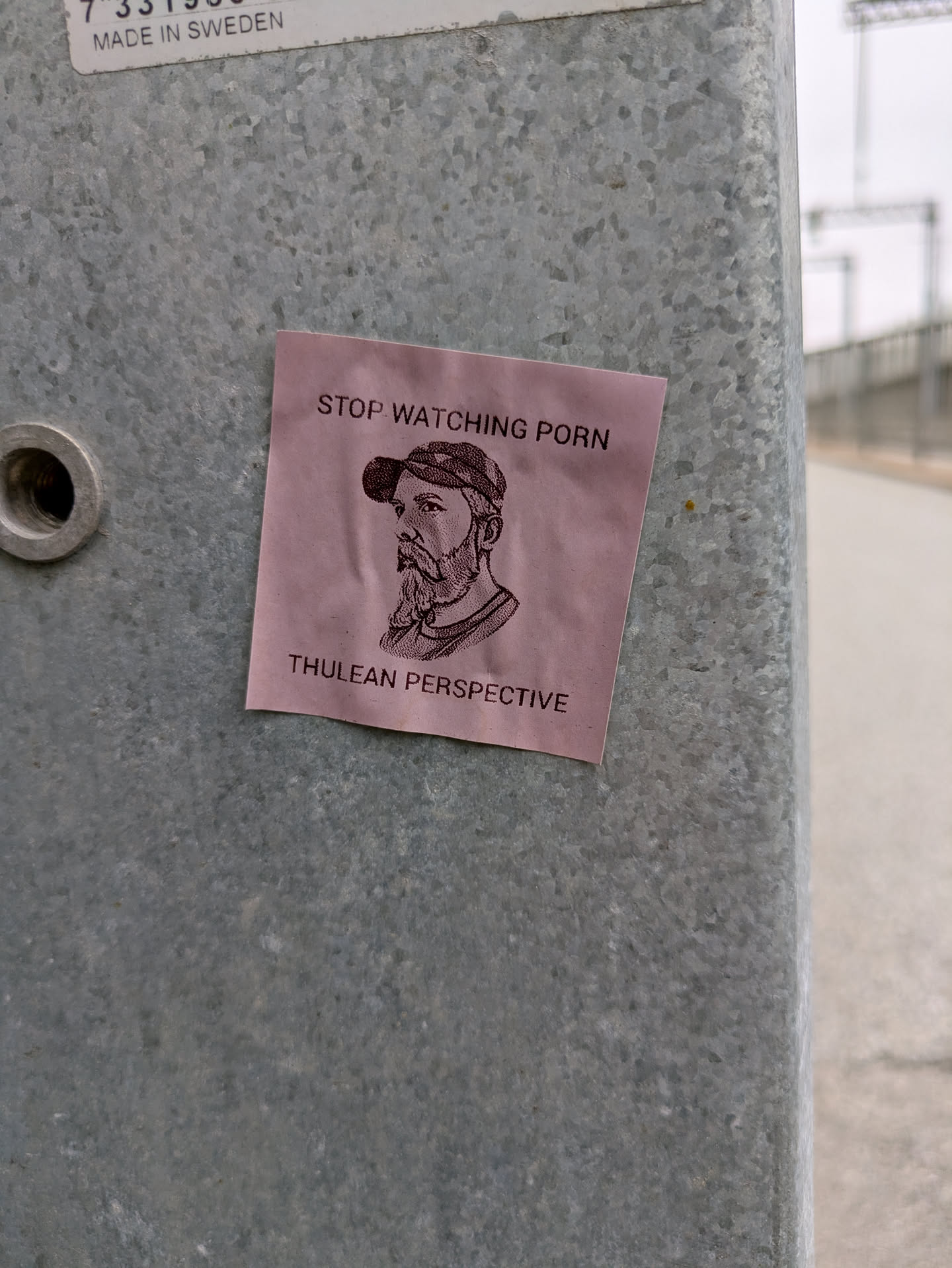 The sticker features a black and white drawing of a bearded man wearing a cap. The background color of the sticker is pink. The sticker is stuck on a metal pole.