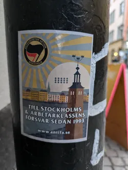 A sticker featuring a sunburst design, a cityscape, and a tower. The text includes 'Antifa' and the year 1993. A website address is also visible.