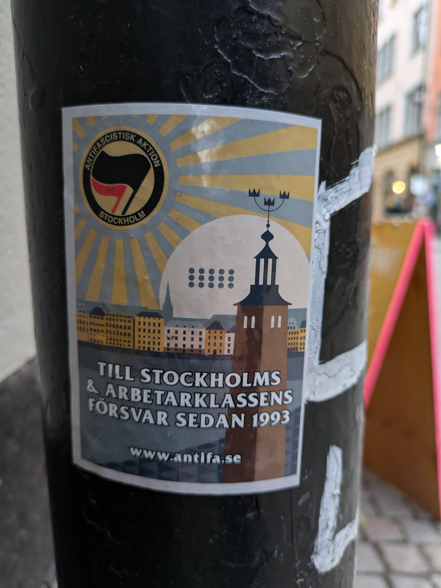 A sticker featuring a sunburst design, a cityscape, and a tower. The text includes 'Antifa' and the year 1993. A website address is also visible.