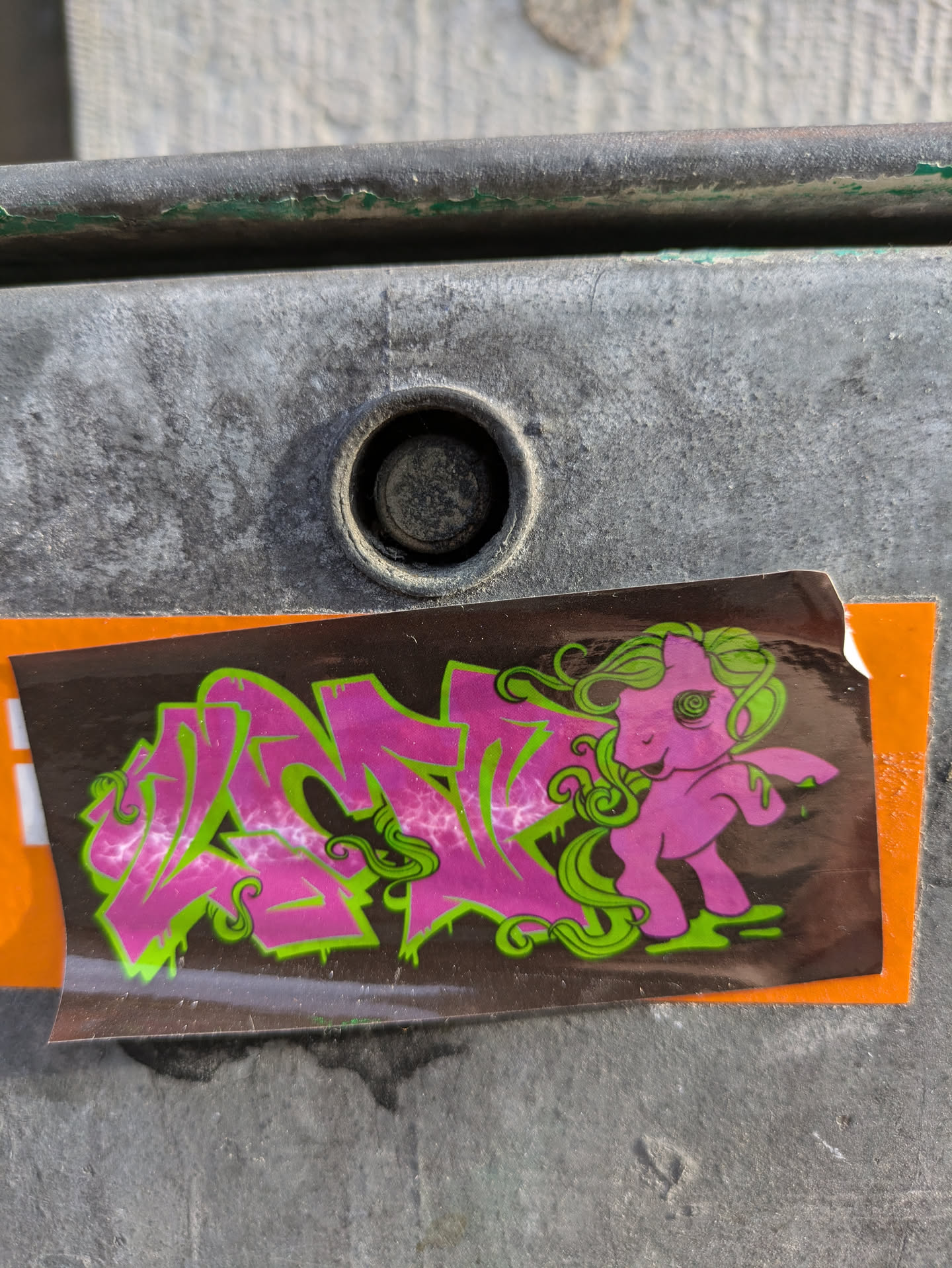 The sticker is dark-colored with pink and green graffiti-style text and a cartoon pony in pink with green hair. It is stuck on a gray metal surface.