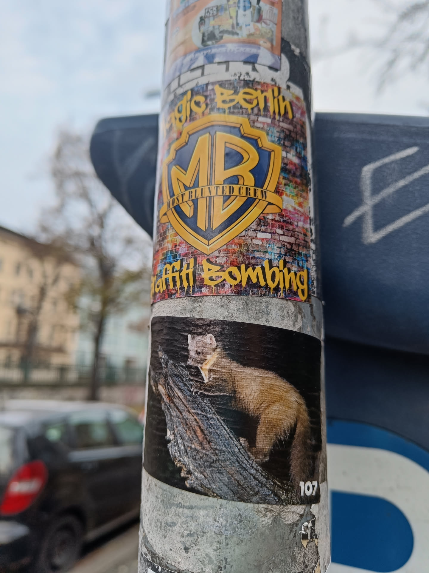 A sticker on a pole in Berlin. The main sticker features the logo for MB Graffiti Berlin Crew. A smaller sticker underneath shows a pine marten. The number 107 is visible on the bottom right of the marten sticker.