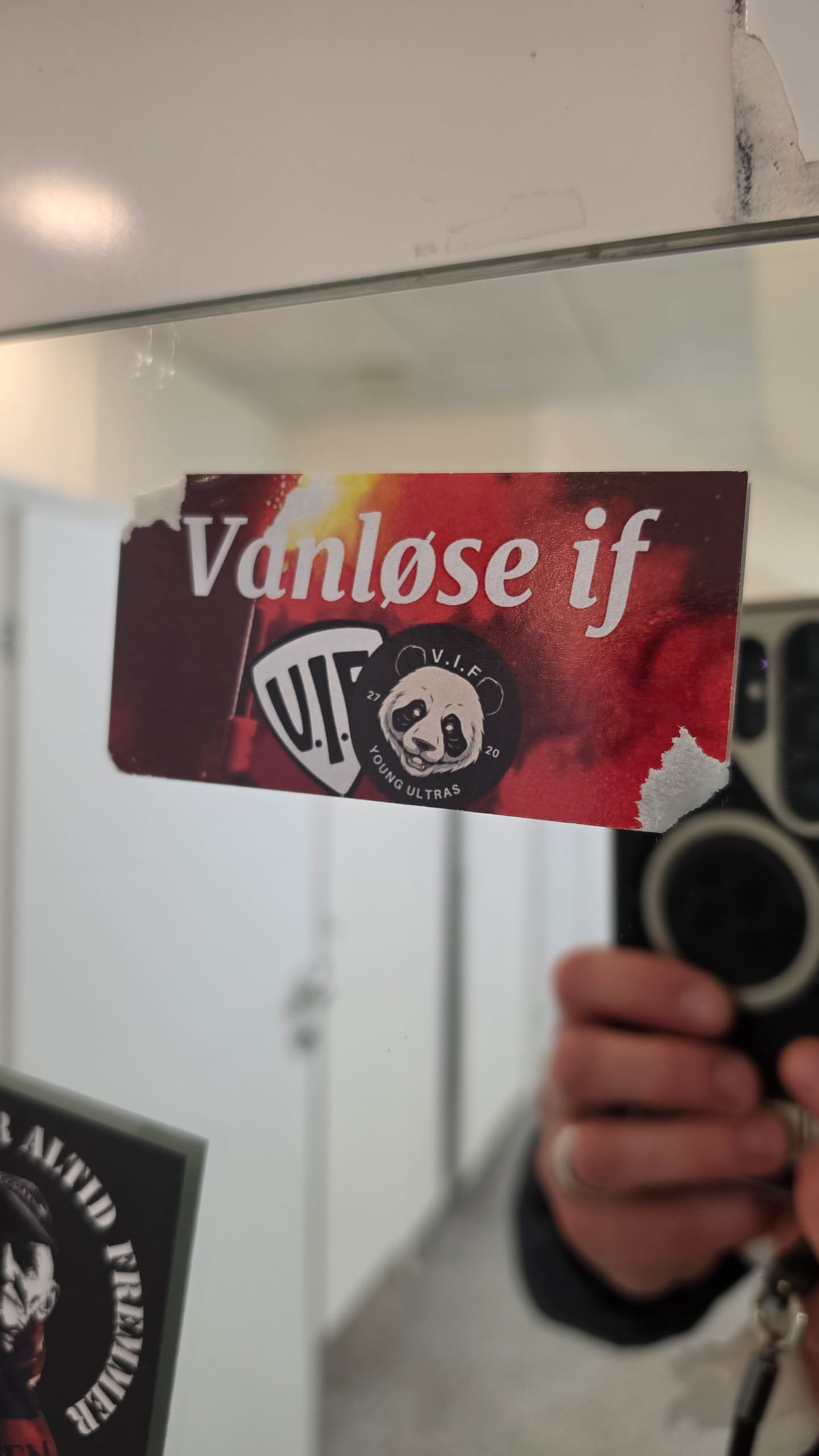 A rectangular sticker placed on a mirror featuring a dark red and smoky background resembling burning pyrotechnics or flares. At the top, the text 'Vanløse if' is written in a white, italic serif font. Beneath the text are two layered emblems: a shield containing the stylized letters 'V.I.F.' and a round black badge showing an illustrated panda face surrounded by the words 'V.I.F.', '27', '20', and 'YOUNG ULTRAS'. The top-left and bottom-right corners of the sticker show signs of wear and peeling.