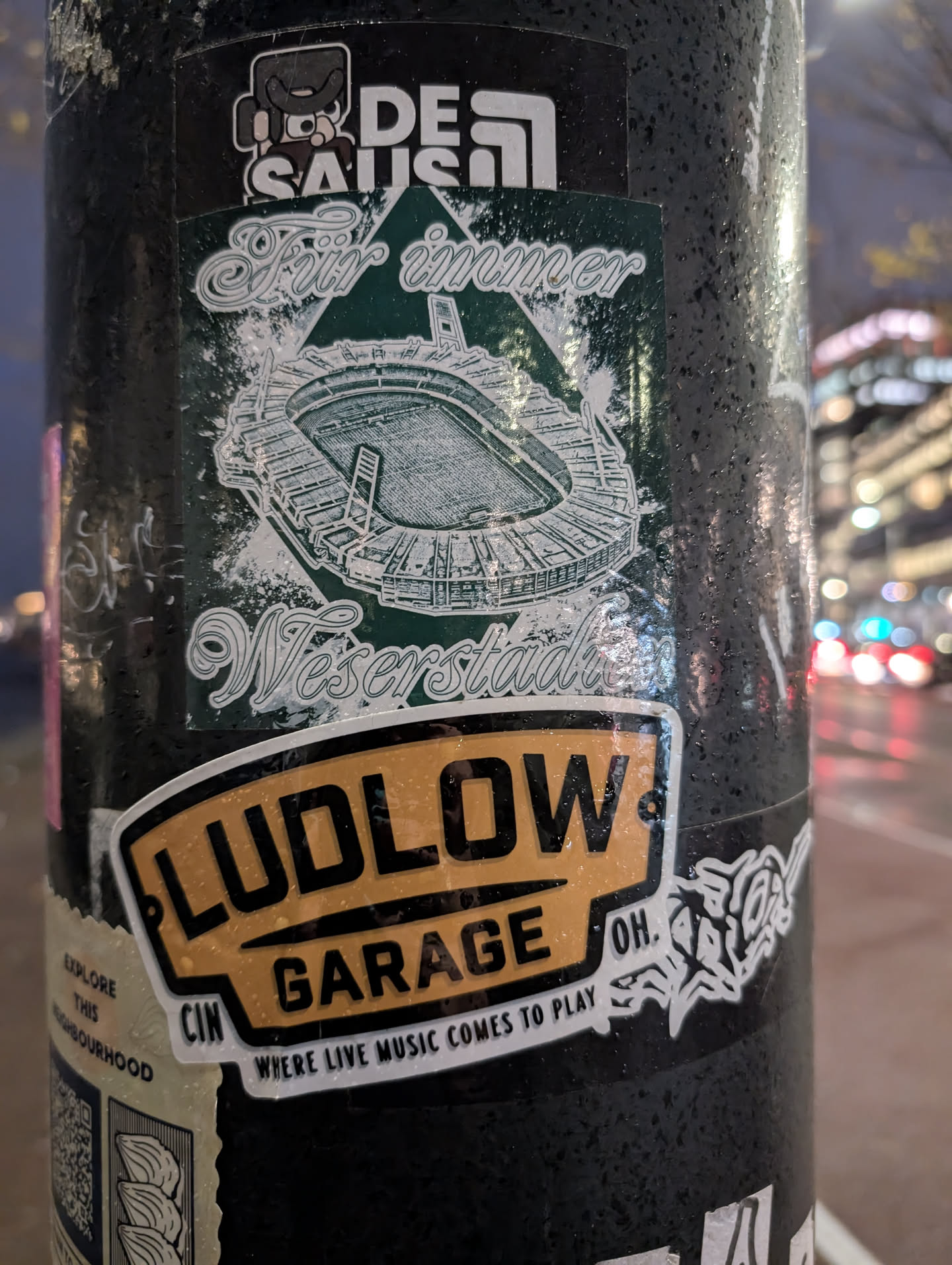 A sticker for Ludlow Garage in Cincinnati, OH.  The sticker features their logo, the text 'Where Live Music Comes to Play', and a smaller sticker underneath featuring a stadium illustration and German text. The smaller sticker has  'Für immer Weserstadion' which translates to 'Forever Weserstadion'. There is also a logo that says De Saus.
