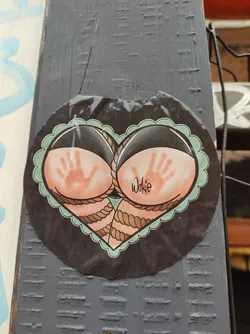 A round sticker in the shape of a heart features two buttocks with handprints on them, tied together with rope. The style is reminiscent of traditional tattoo art. The artist's signature, "Wokie," is visible. The sticker is affixed to a dark gray wooden surface.