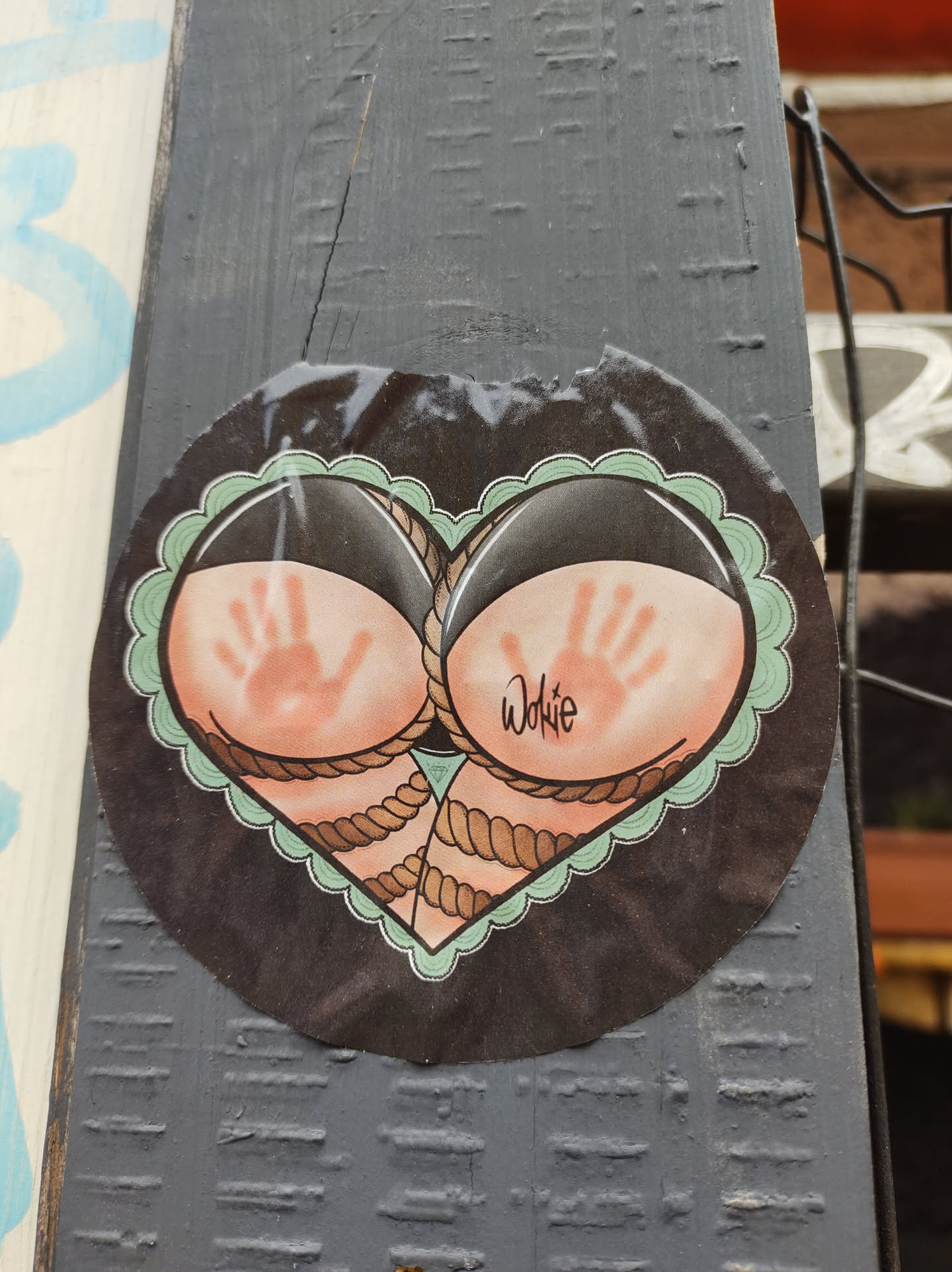 A round sticker in the shape of a heart features two buttocks with handprints on them, tied together with rope. The style is reminiscent of traditional tattoo art. The artist's signature, "Wokie," is visible. The sticker is affixed to a dark gray wooden surface.