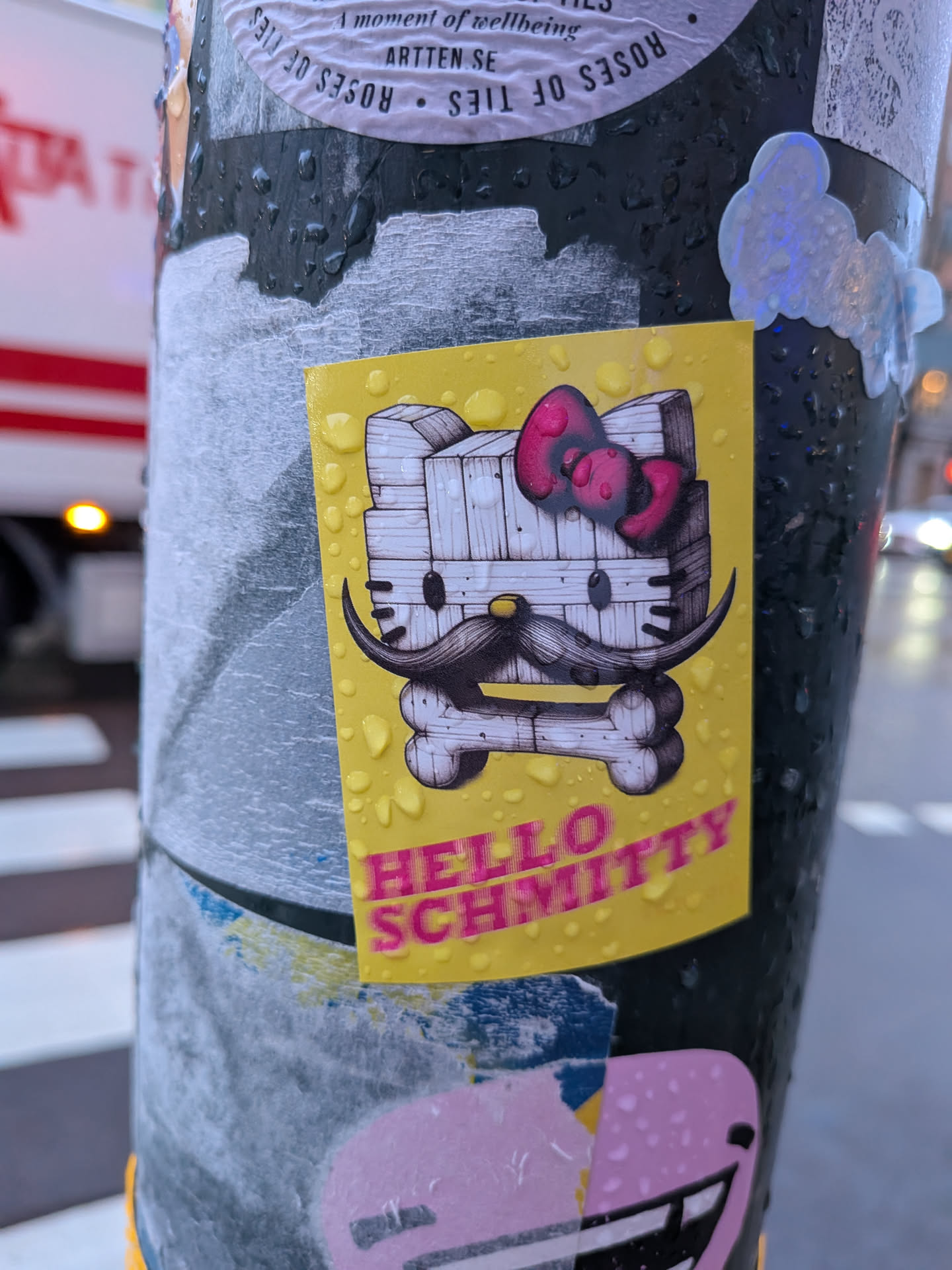 A vertical rectangular sticker with a bright yellow background, covered in water droplets. It features a stylized character based on Hello Kitty, depicted as if built from rough white wooden planks. The character wears a dark pink bow on her left ear, has small dark eyes, a yellow button nose, and a large black handlebar mustache curling upwards. A white wooden bone rests beneath her chin. Printed in bold pink capital letters at the bottom is the parody text 'HELLO SCHMITTY'.