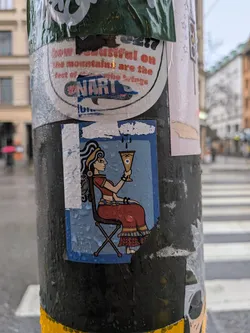 The central sticker features a stylized illustration of a woman seated in profile, facing right. She has dark, wavy hair adorned with a yellow headband and a prominent blue earring. Her skin tone is light brown. She wears a long, flowing red dress with intricate yellow and blue patterns on the hem and waist, and a yellow bracelet on her left wrist. She is seated on a simple brown folding chair with an 'X' shaped base. In her right hand, she holds a golden goblet with a distinct blue eye symbol in its center, from which dark droplets appear to fall. The background of the sticker is a solid light blue, framed by a darker blue border. The sticker is rectangular, showing visible wear, creases, and water droplets on its surface, indicating it is a physical sticker adhered to a pole.