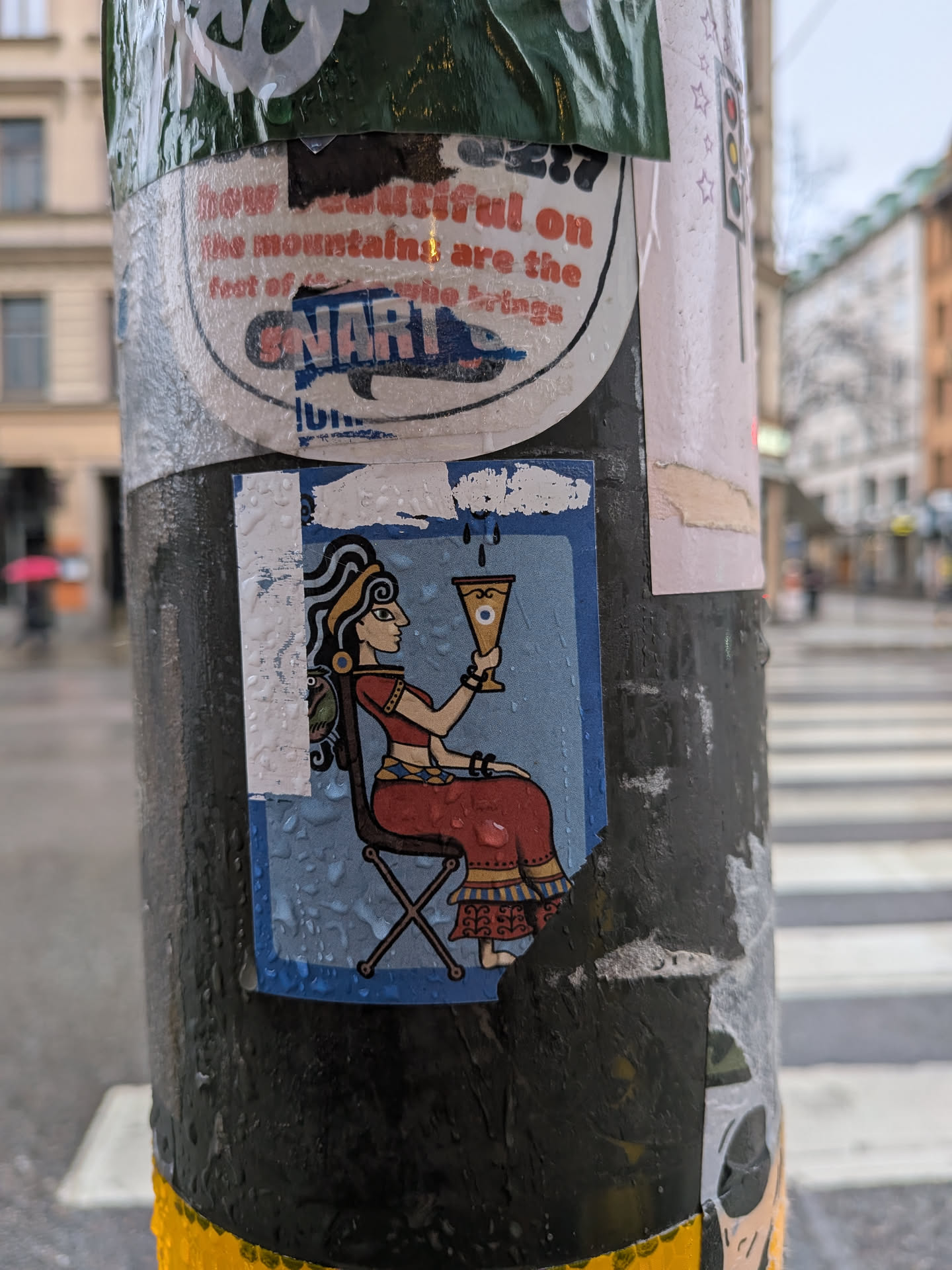 The central sticker features a stylized illustration of a woman seated in profile, facing right. She has dark, wavy hair adorned with a yellow headband and a prominent blue earring. Her skin tone is light brown. She wears a long, flowing red dress with intricate yellow and blue patterns on the hem and waist, and a yellow bracelet on her left wrist. She is seated on a simple brown folding chair with an 'X' shaped base. In her right hand, she holds a golden goblet with a distinct blue eye symbol in its center, from which dark droplets appear to fall. The background of the sticker is a solid light blue, framed by a darker blue border. The sticker is rectangular, showing visible wear, creases, and water droplets on its surface, indicating it is a physical sticker adhered to a pole.