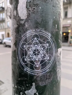 A round, transparent sticker featuring a geometric design is affixed to a textured, dark-colored pole. The sticker promotes Stockholm Tattooing and has an Instagram handle for Andreas Engblom.