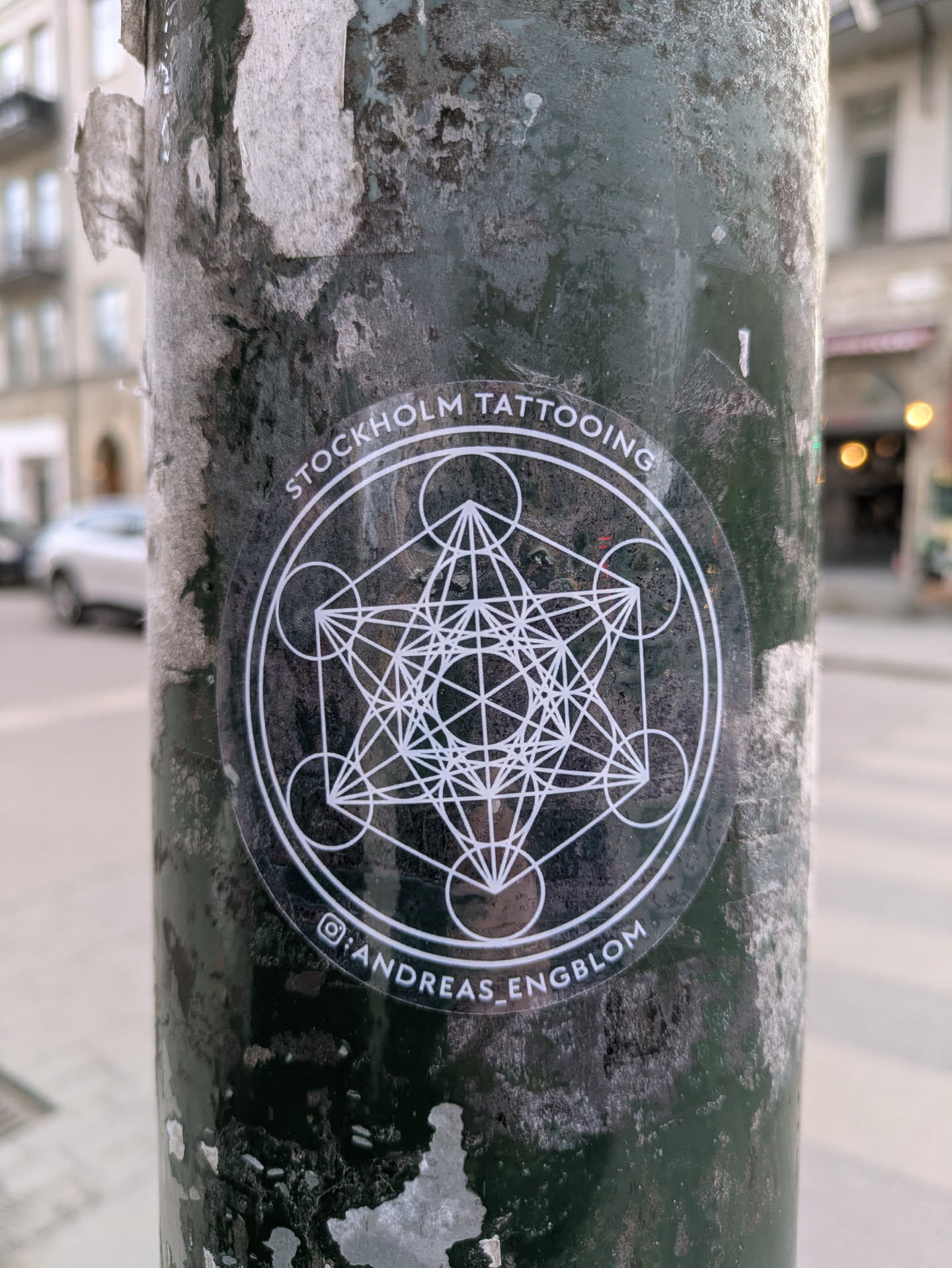 A round, transparent sticker featuring a geometric design is affixed to a textured, dark-colored pole. The sticker promotes Stockholm Tattooing and has an Instagram handle for Andreas Engblom.