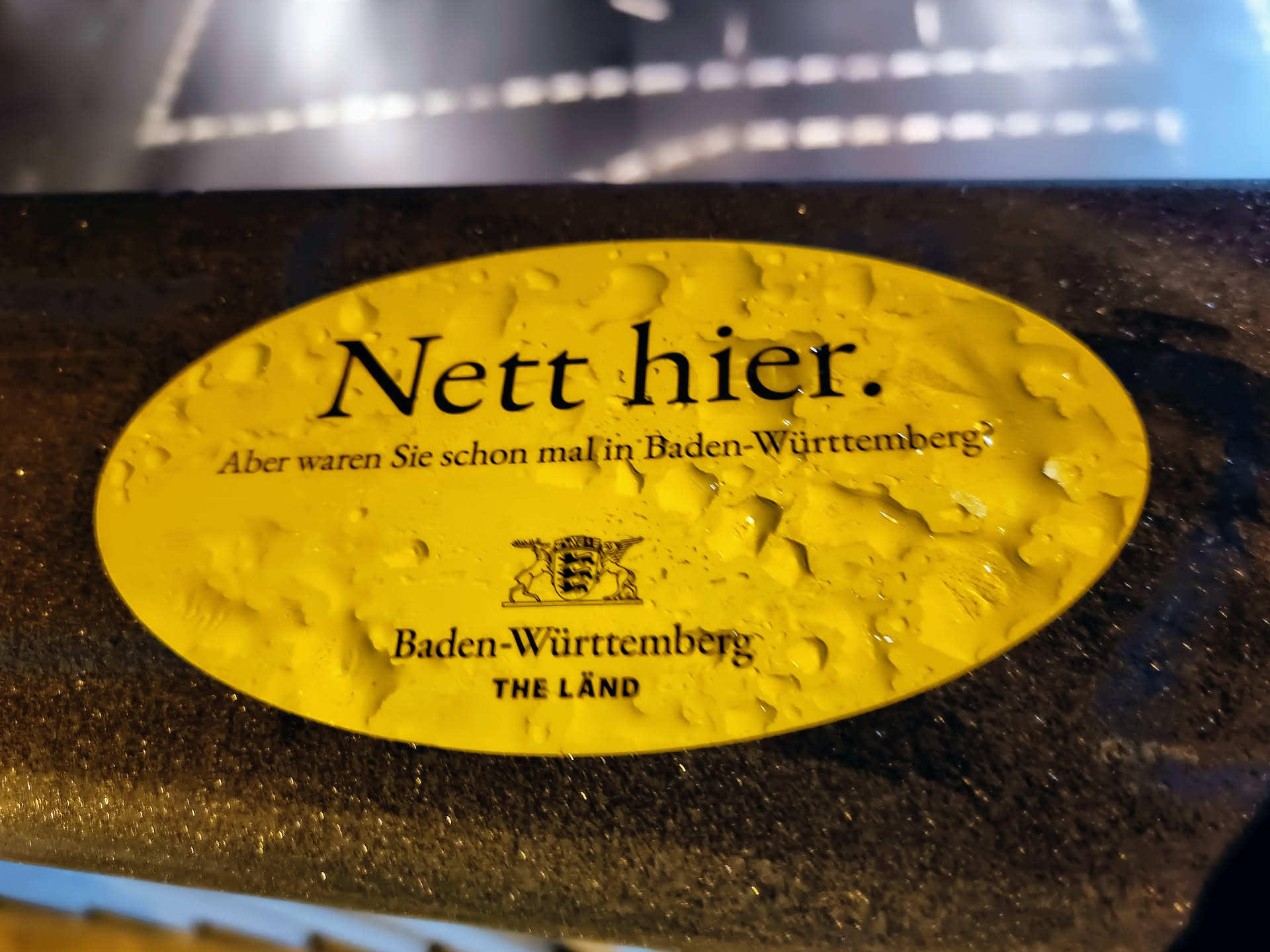 Yellow oval sticker with the text "Nett hier." (Nice here.) and  "Aber waren Sie schon mal in Baden-Württemberg?" (But have you ever been to Baden-Württemberg?). It also shows the coat of arms of Baden-Württemberg and the text "Baden-Württemberg THE LÄND". The sticker seems to be attached to a dark surface and has water droplets on it.
