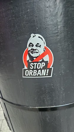 The sticker depicts a black and white portrait of Viktor Orban, the Prime Minister of Hungary, with a red prohibition sign imposed on the face. Below the portrait, the sticker reads "STOP ORBAN!" in bold black letters on a white background. The sticker is affixed to a black surface.