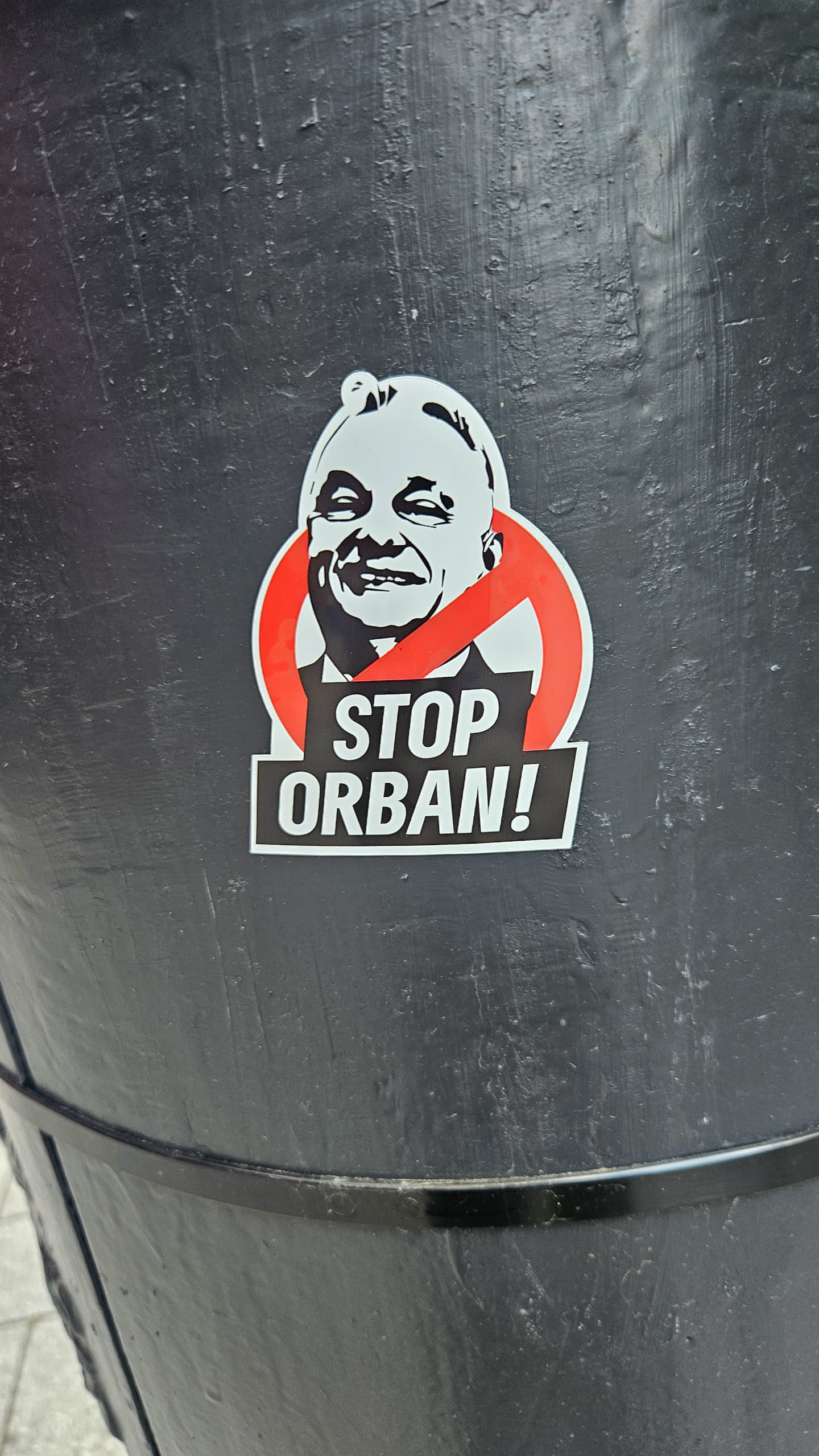 The sticker depicts a black and white portrait of Viktor Orban, the Prime Minister of Hungary, with a red prohibition sign imposed on the face. Below the portrait, the sticker reads "STOP ORBAN!" in bold black letters on a white background. The sticker is affixed to a black surface.