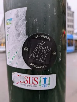 The sticker is round and black, featuring the text "HALVNORSK" at the top and bottom. An abstract, stylized logo is placed in the center of the sticker. There are Spotify logos on either side of the central logo. The sticker is affixed to a green pole with other stickers.