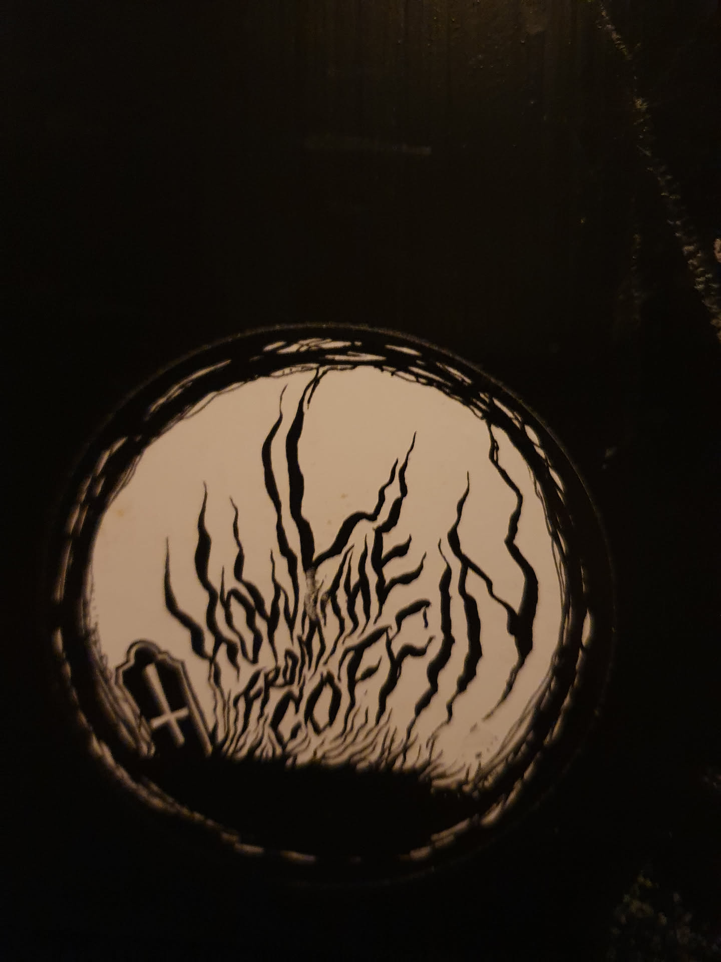 A round sticker featuring a dark, gothic design. The central motif appears to be a gravestone with flames or vines emanating from it.  The text reads "FROM THE COFFIN". The style is reminiscent of darkwave or death metal aesthetics.