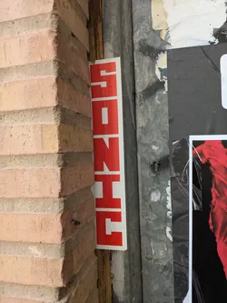The sticker is red and white and vertically oriented, it spells the word 'SONHE' in Portuguese, meaning 'dream'. It's placed between the corner of a brick wall and a weathered metal surface with other posters.