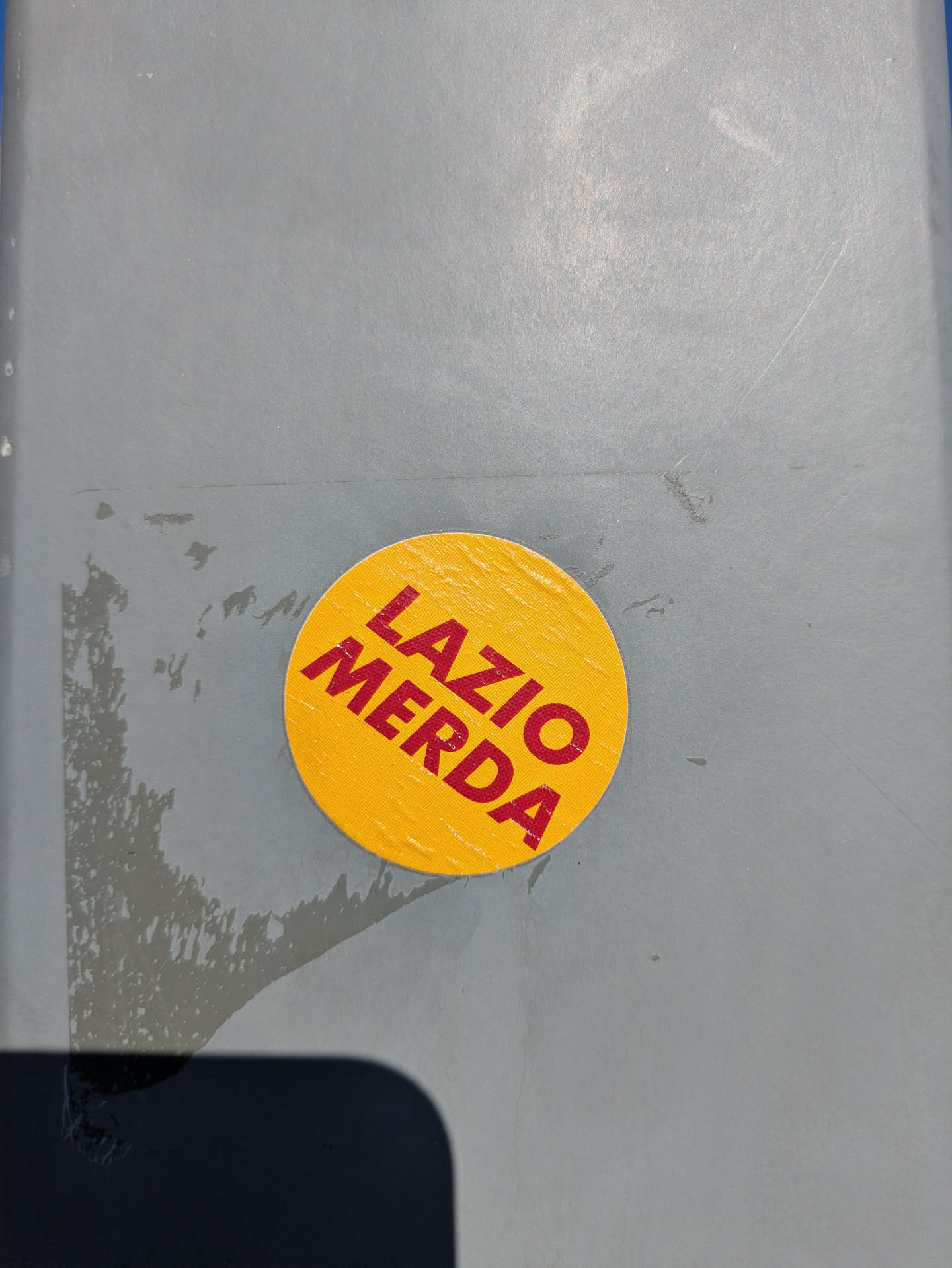 This is a round, yellow sticker with the words "LAZIO MERDA" printed in red. The sticker is stuck on a gray surface, possibly a wall or door. The text is in bold and slightly tilted.