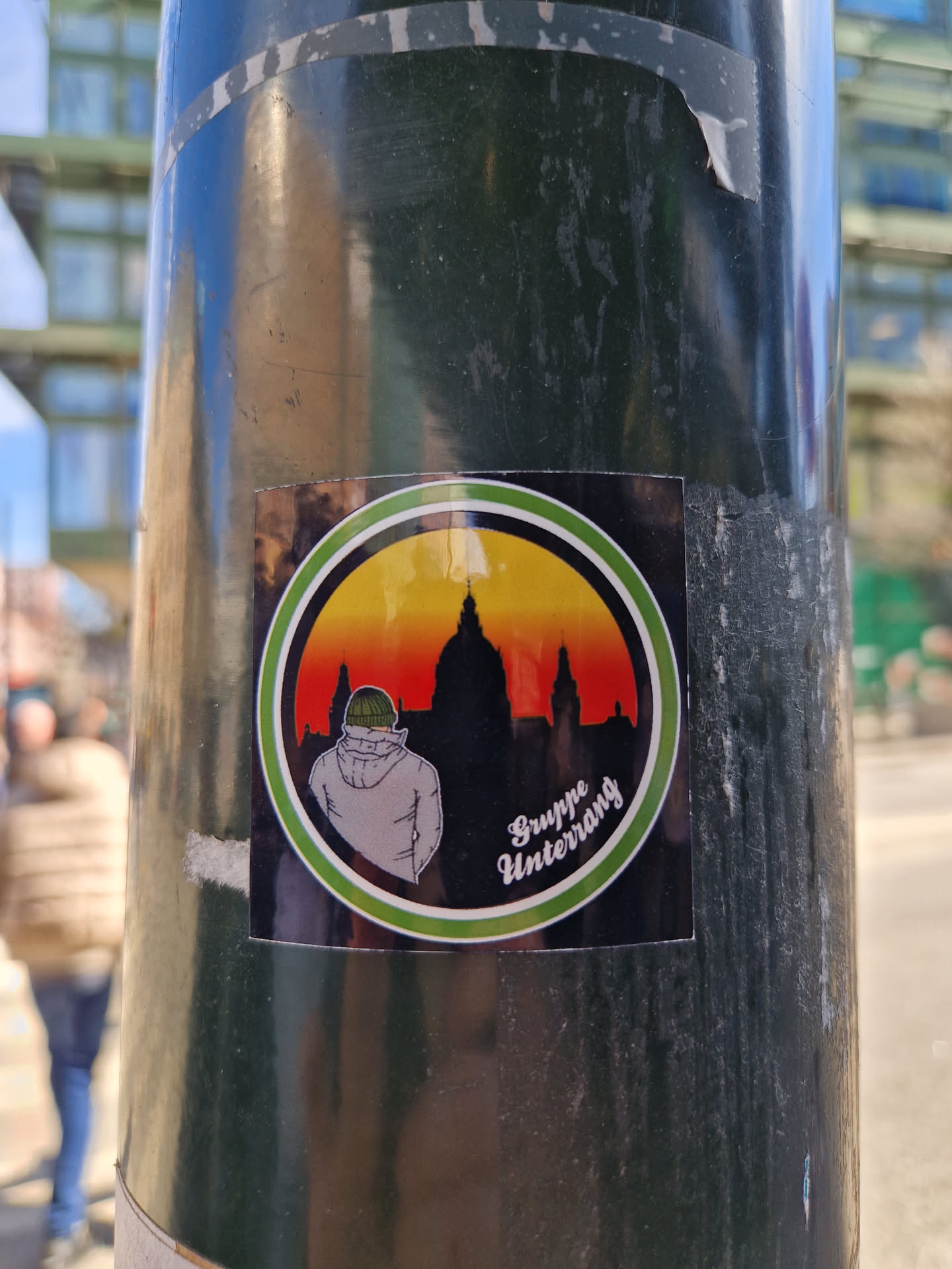 This round sticker shows a stylized person from behind, seemingly wearing a beanie or helmet, set against a vibrant sunset and a silhouette of what looks like a European city skyline, possibly including a dome-like structure. The text at the bottom reads 'Gruppe Untergang' in a stylized font.
