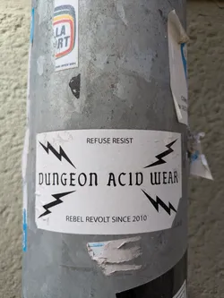 A white sticker on a pole with the text 'DUNGEON ACID WEAR, REFUSE RESIST, REBEL REVOLT SINCE 2010' in black, with lightning bolts around the brand name. The text is blocky, and the aesthetic seems punk or alternative.