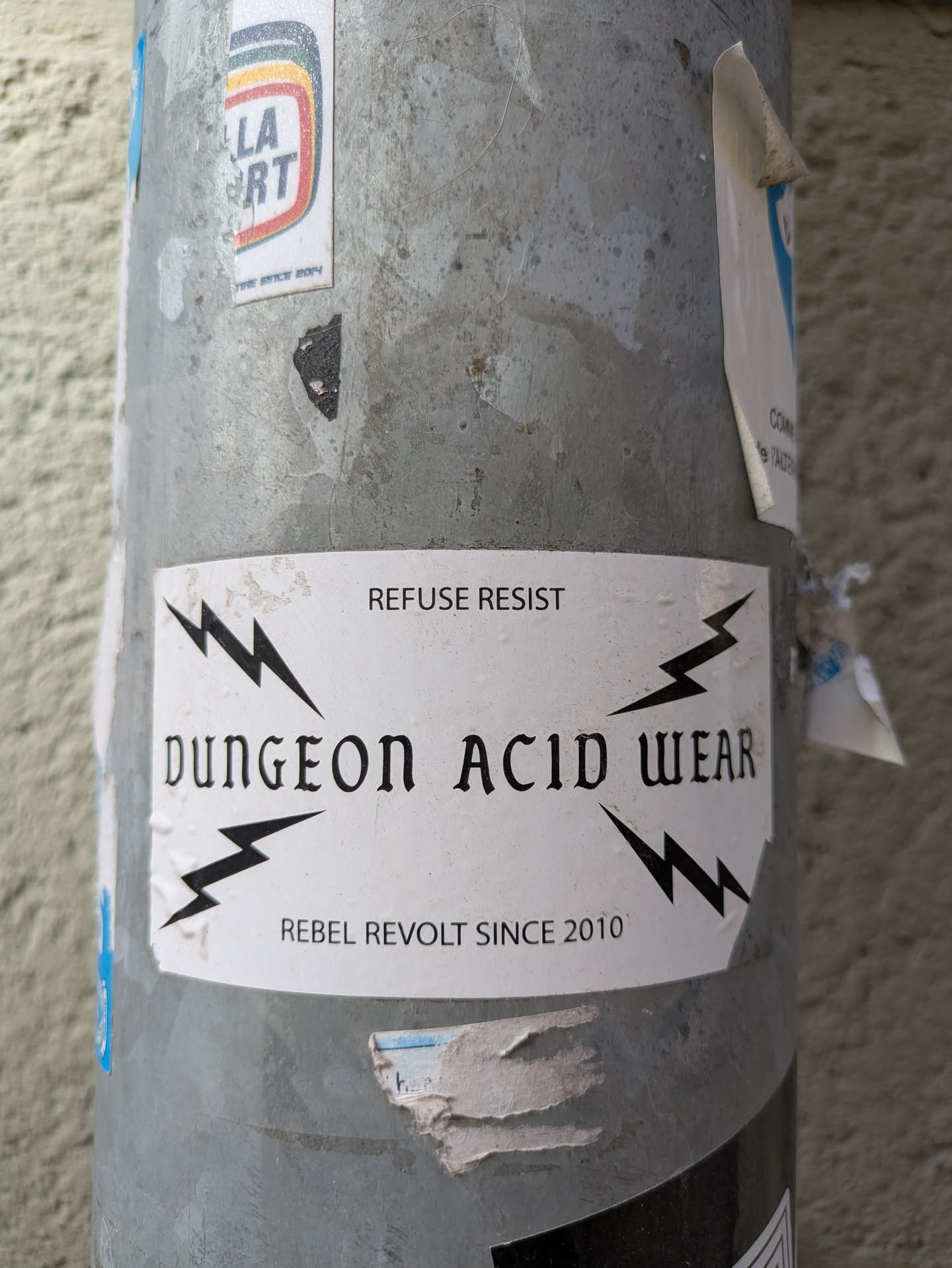 A white sticker on a pole with the text 'DUNGEON ACID WEAR, REFUSE RESIST, REBEL REVOLT SINCE 2010' in black, with lightning bolts around the brand name. The text is blocky, and the aesthetic seems punk or alternative.