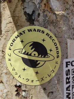 Yellow round sticker with a black vinyl record orbiting a planet. The text says "Format Wars Records" in English and  "フォーマット・ウォーズ・レコード" in Japanese. 