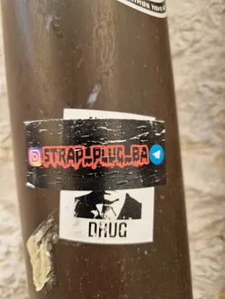 A sticker with a stylized image of a man in a suit and tie, and the word "DRUG" underneath.  The top of the sticker features a social media handle, @STRAP_PLUG_BA, with Instagram and Telegram icons.