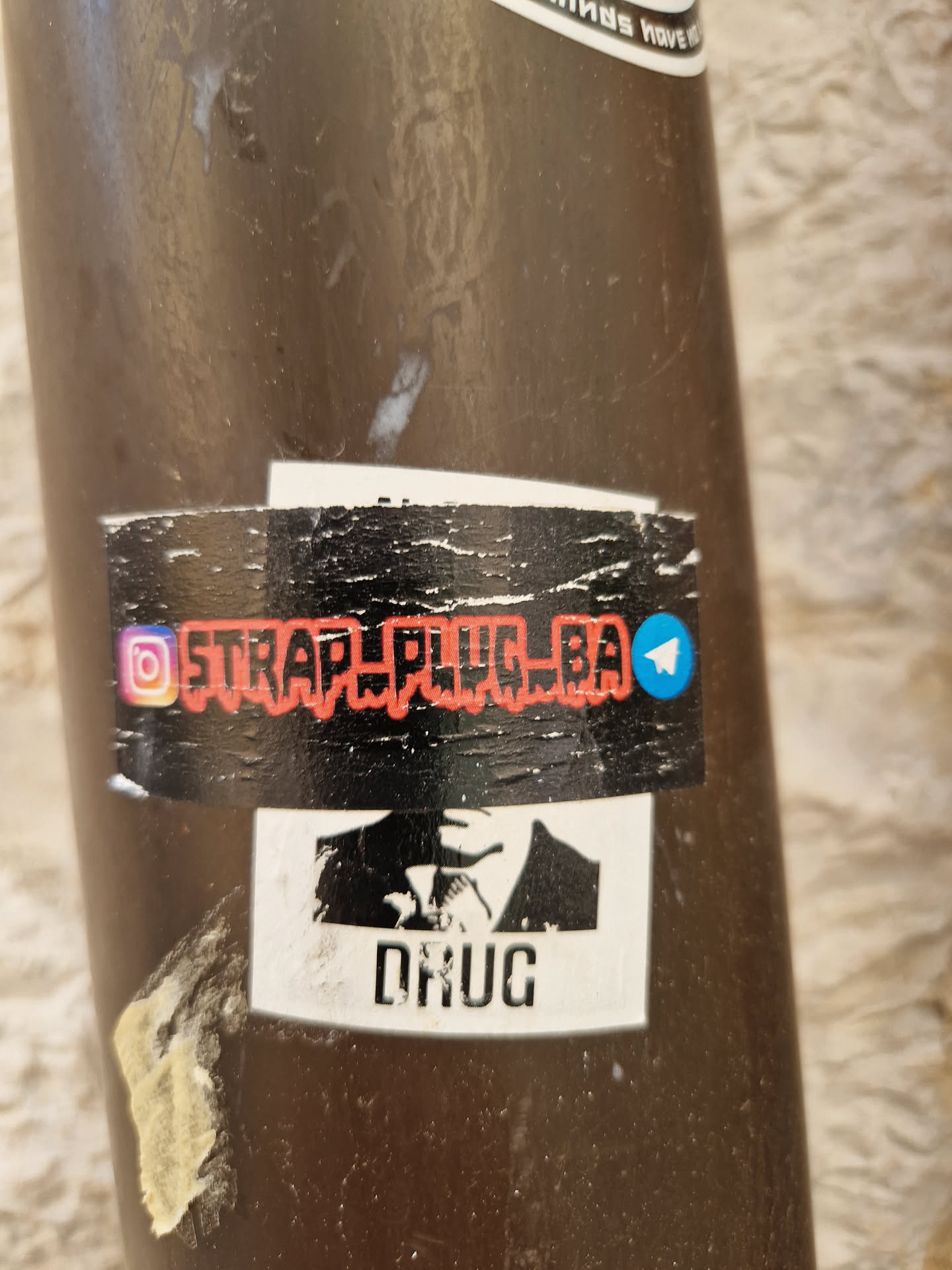 A sticker with a stylized image of a man in a suit and tie, and the word "DRUG" underneath.  The top of the sticker features a social media handle, @STRAP_PLUG_BA, with Instagram and Telegram icons.