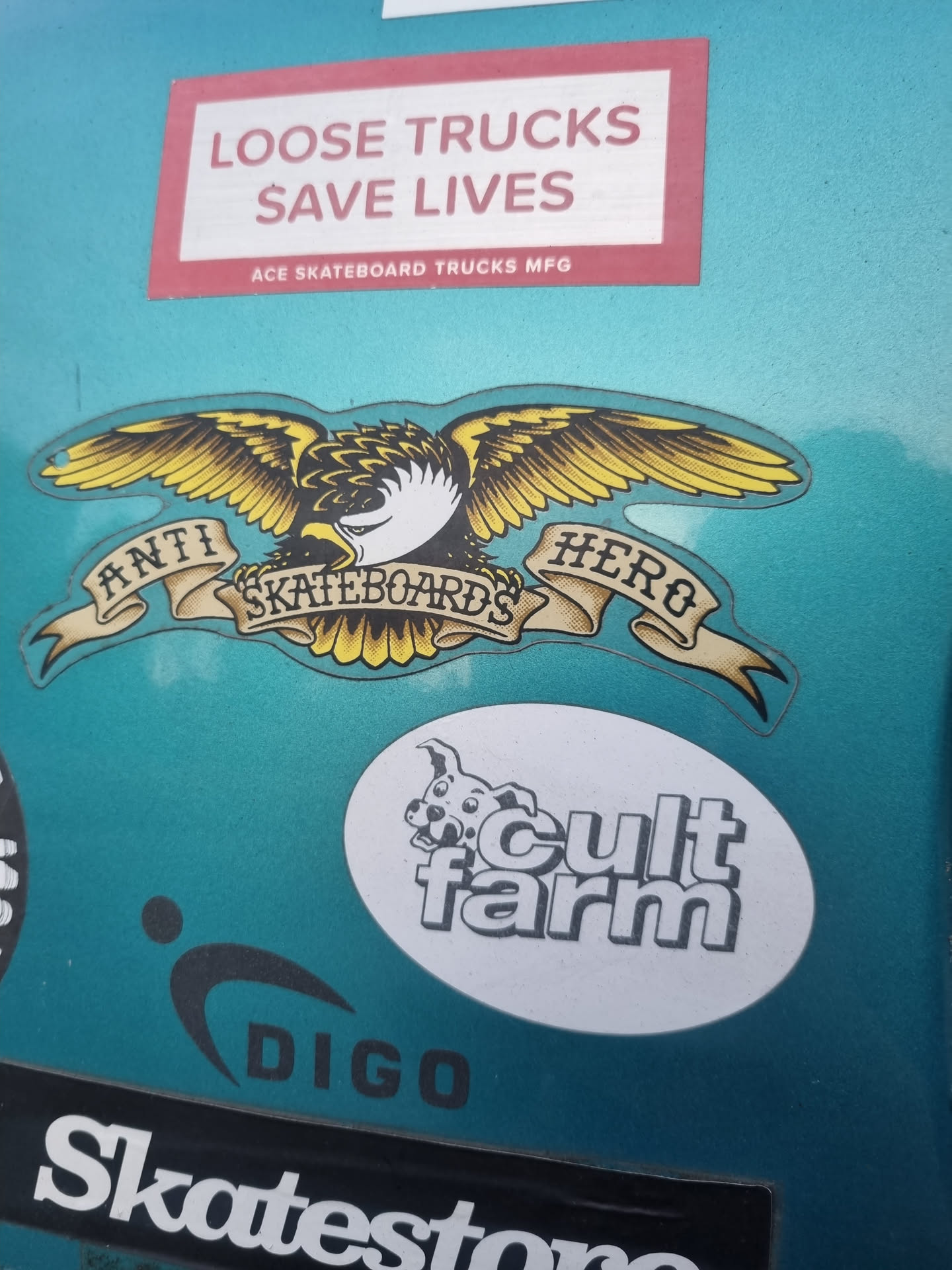 A teal surface with various skateboarding brand stickers. The main focus is an Anti Hero Skateboards sticker featuring their iconic eagle logo with the words "Anti Hero Skateboards" written on a banner underneath the eagle. Other stickers include "Loose Trucks Save Lives", "Cult Farm", and "Digo".