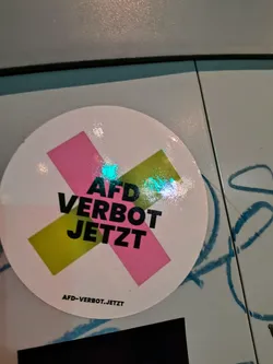 A round sticker with a pink and yellow X design, overlaid with the words "AFD VERBOT JETZT" in bold black letters. The website "AFD-VERBOT.JETZT" is printed at the bottom of the sticker.