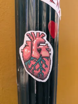 A pixelated anatomical heart sticker is placed on a black, ridged pole with a yellow wall in the background. The sticker is mostly pink, red and teal. It looks like a pixel art version of a real heart.