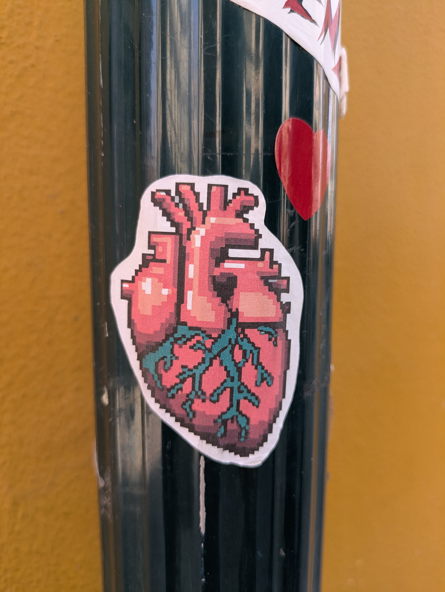A pixelated anatomical heart sticker is placed on a black, ridged pole with a yellow wall in the background. The sticker is mostly pink, red and teal. It looks like a pixel art version of a real heart.