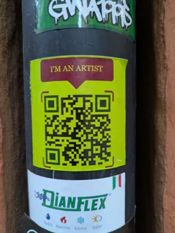A yellow QR code sticker with the text "I'M AN ARTIST" on a dark maroon background. The sticker is placed on a dark gray surface, which seems to be a pipe or similar cylindrical object. There are other stickers around it.