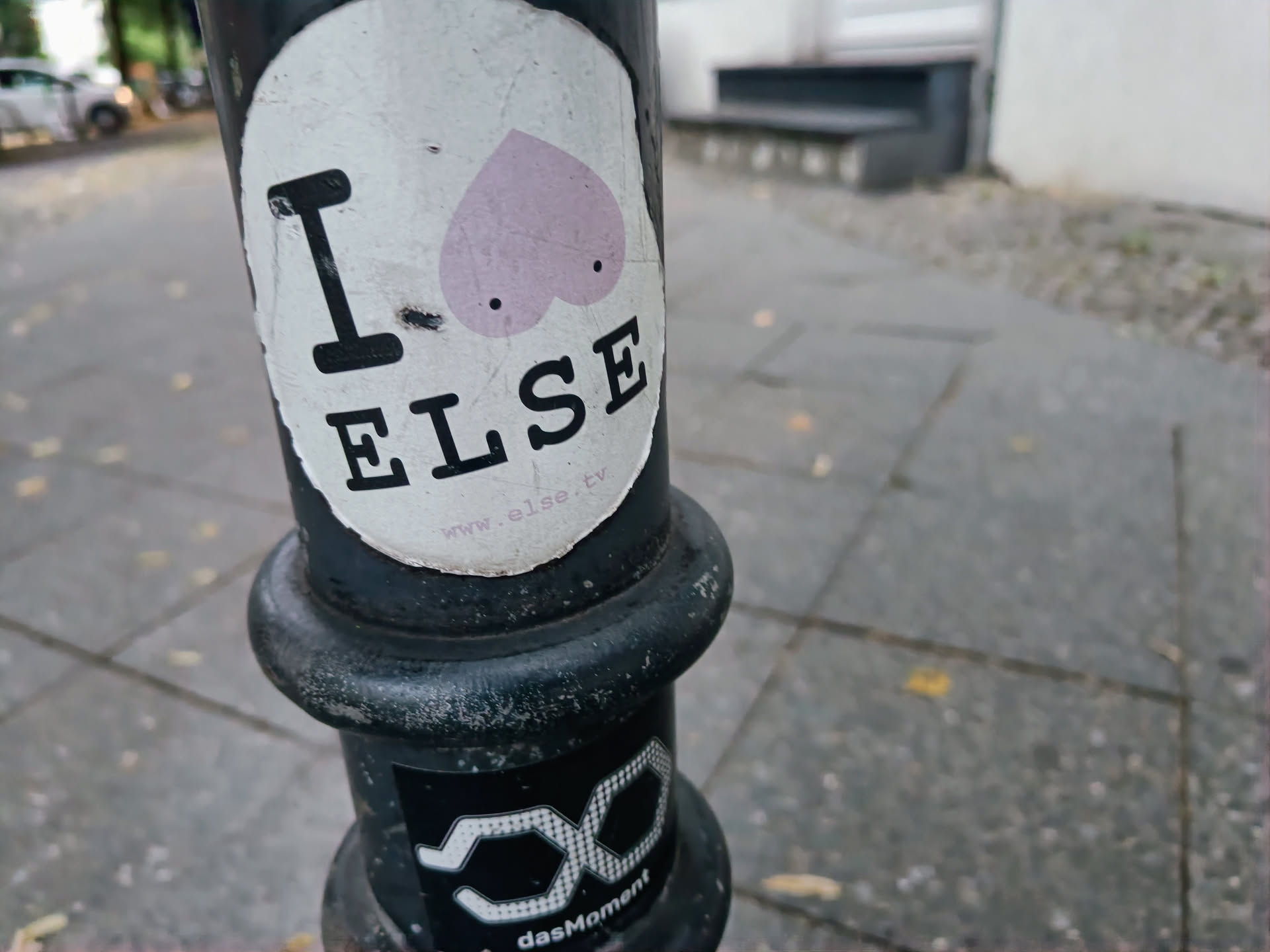 A sticker with a minimalist design featuring the text "I \u2764 ELSE" and a website address. The heart includes two small circles that resemble eyes. The sticker is affixed to a lamppost.