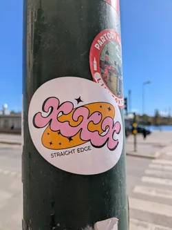 A round sticker with a pink, stylized, bubble letter design over a yellow background dotted with small stars.  The words "STRAIGHT EDGE" are printed beneath the design.