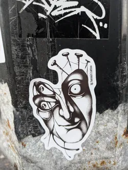 A black and white sticker depicting a surreal image. A human face appears to be merging with a butterfly, with several nails piercing the top of the head. The style is reminiscent of dark art or surrealism.  The sticker has the Instagram handle '@ig.bartolo.tattoo' printed on it.
