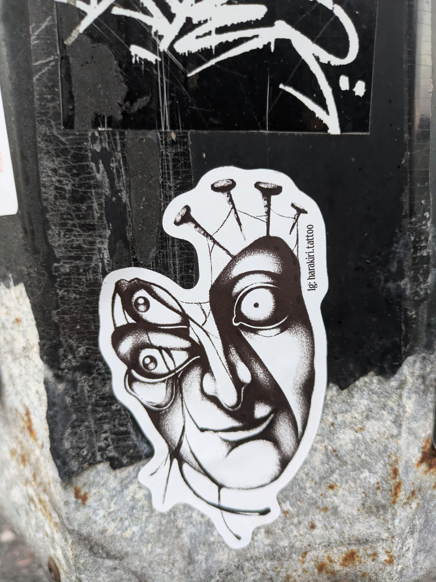 A black and white sticker depicting a surreal image. A human face appears to be merging with a butterfly, with several nails piercing the top of the head. The style is reminiscent of dark art or surrealism.  The sticker has the Instagram handle '@ig.bartolo.tattoo' printed on it.