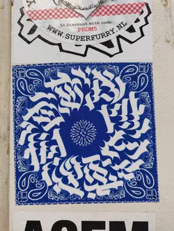 A square sticker with a blue bandana print as background. The main design is an abstract pattern in white, featuring stylized lettering or symbols arranged in a circular composition.  A smaller floral element is in the center of the circle. The style is reminiscent of graffiti or street art.