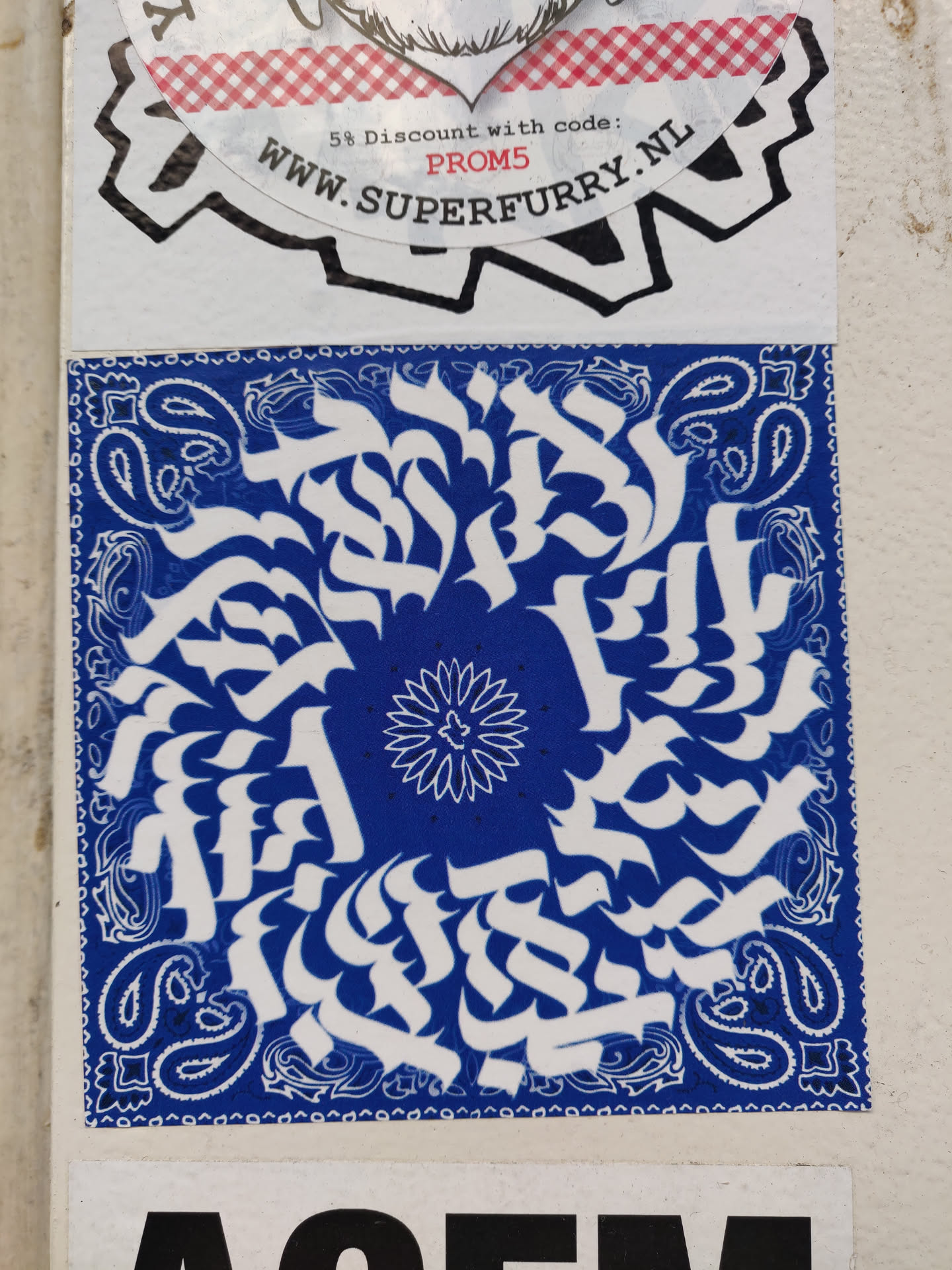 A square sticker with a blue bandana print as background. The main design is an abstract pattern in white, featuring stylized lettering or symbols arranged in a circular composition.  A smaller floral element is in the center of the circle. The style is reminiscent of graffiti or street art.