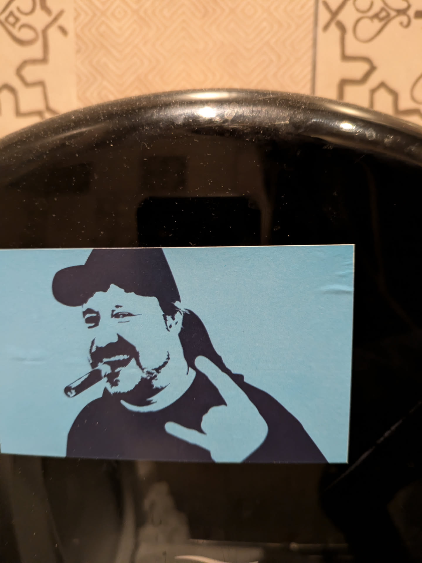 This sticker shows a man with a cap, a cigar in his mouth, and a rock and roll hand gesture. The style is reminiscent of a stencil or screen print, with bold outlines and flat colors. The background is light blue, contrasting with the dark blue figure. The sticker appears to be applied to a dark surface.