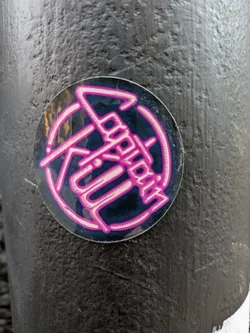Round sticker with a dark background and pink logo. The logo appears to say "Logokill" in a stylized font. 