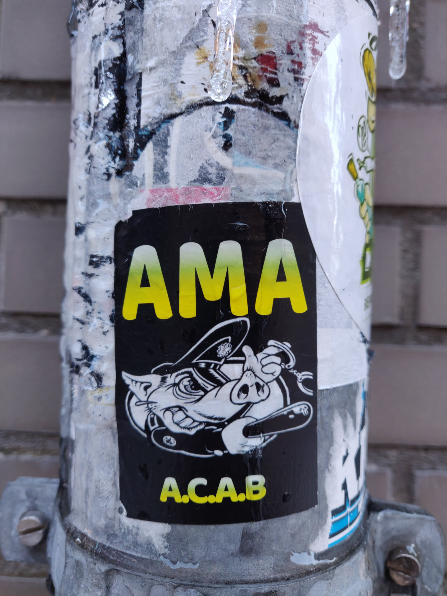 A black and yellow sticker with the text "AMA" and "A.C.A.B." It features an illustration of a pig wearing a police uniform and holding handcuffs. The sticker is affixed to a metal pole, which is covered in other stickers and grime. There is ice forming on the pole.