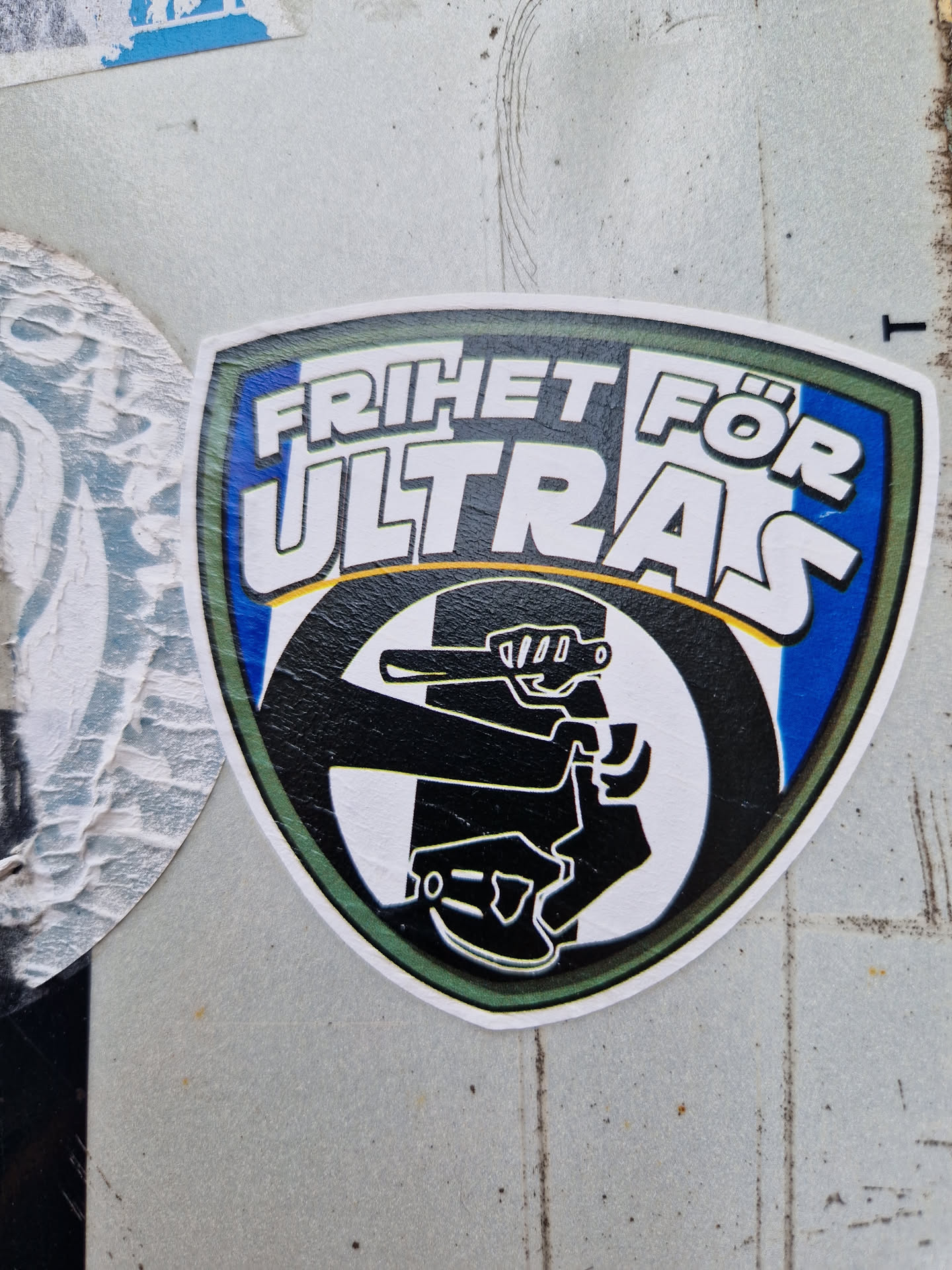 A sticker with a shield shape and the text "Frihet för Ultras" (Freedom for Ultras) in bold letters. It features a stylized graphic of a person holding what seems to be a gun or a similar object. The color scheme consists of black, white, blue, and green.