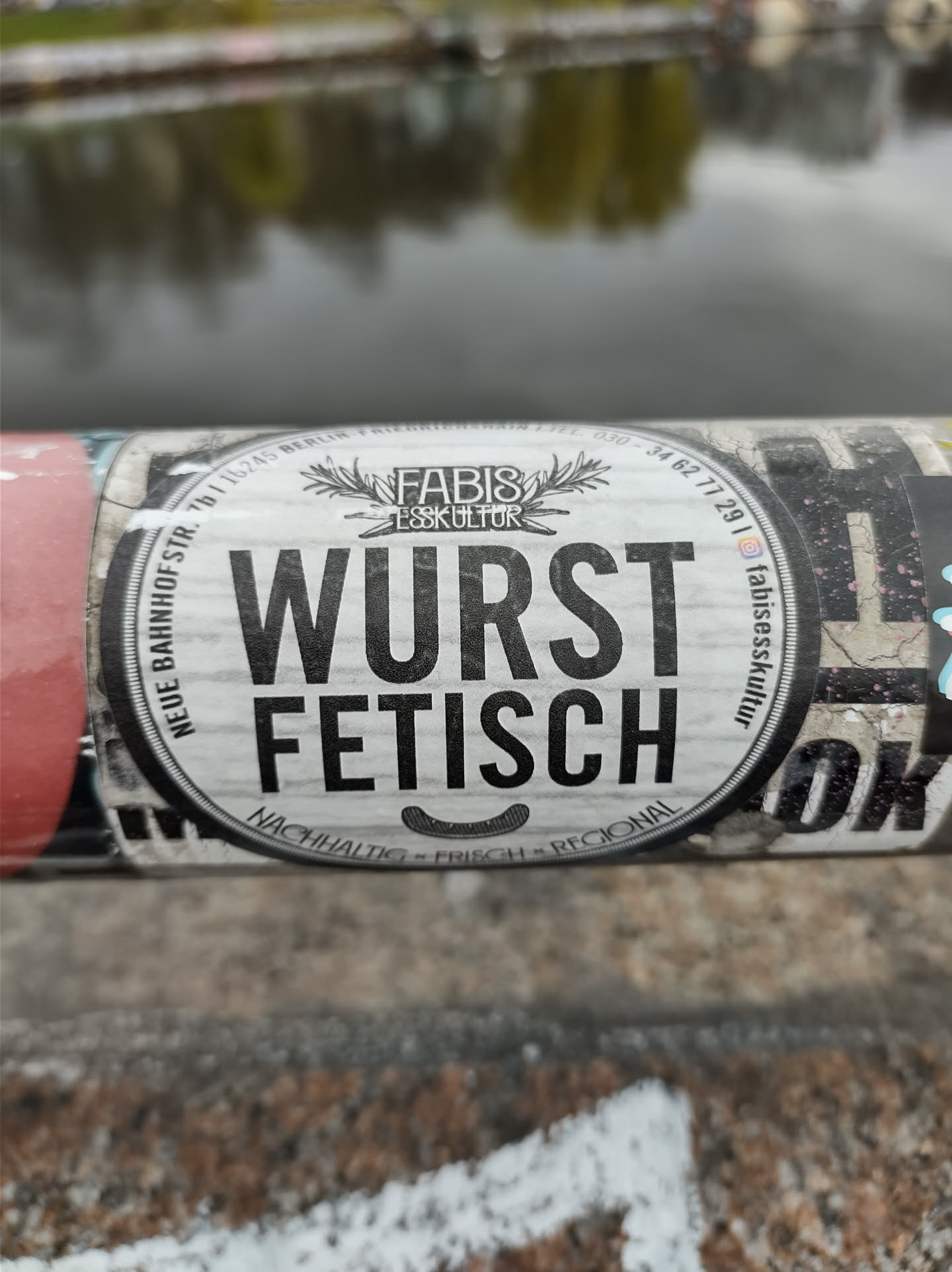 Round sticker with the text "Wurst Fetisch" in the center. It also contains the logo of Fabis Esskultur and some additional text in a smaller font. The sticker is placed on a pole.