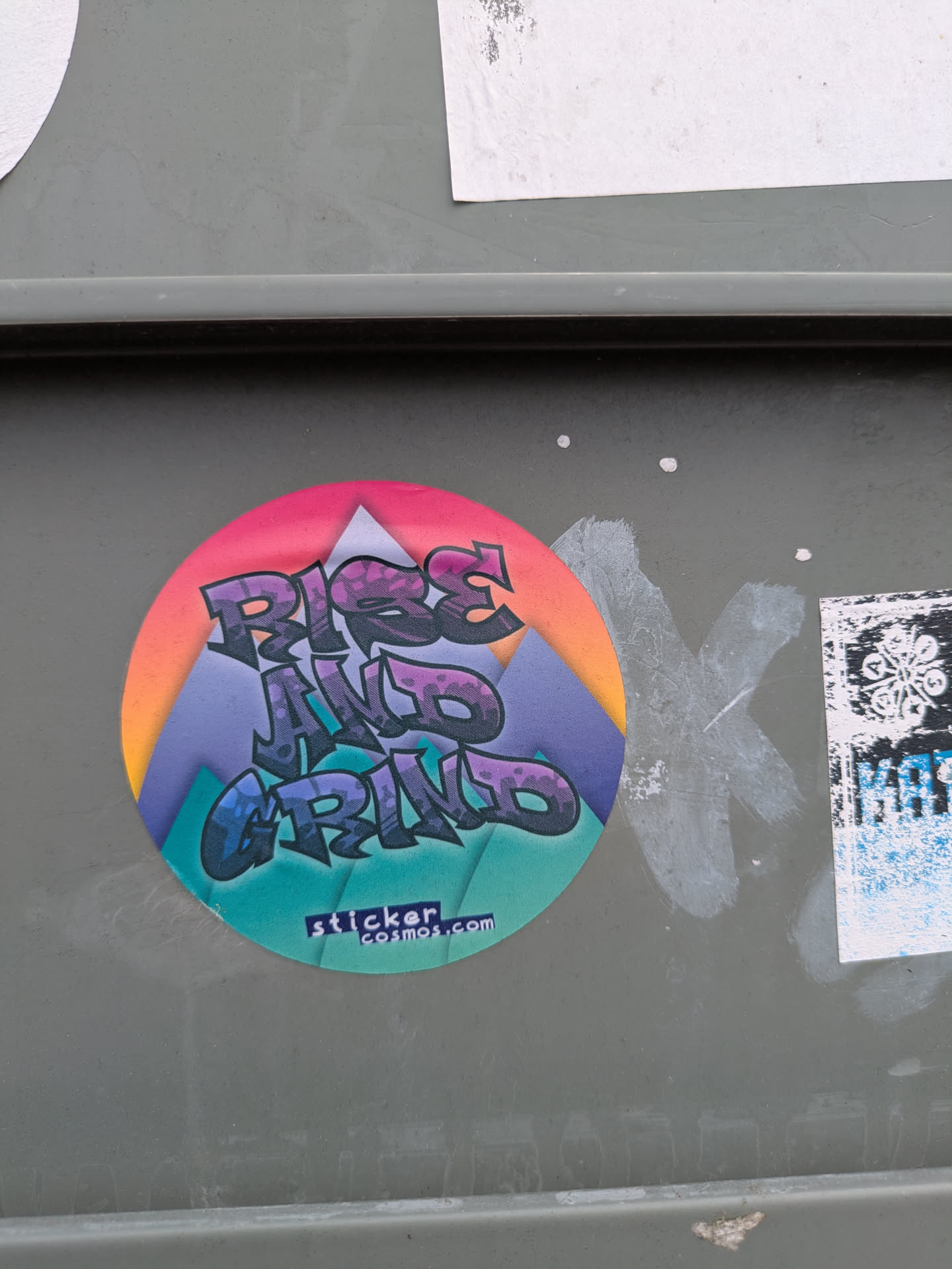 A round sticker with the words "Rise and Grind" in stylized graffiti font, set against a mountain range illustration. The sticker has a vibrant color gradient, with pink and orange at the top fading to blue and green at the bottom. The URL stickercosmos.com is printed at the bottom.