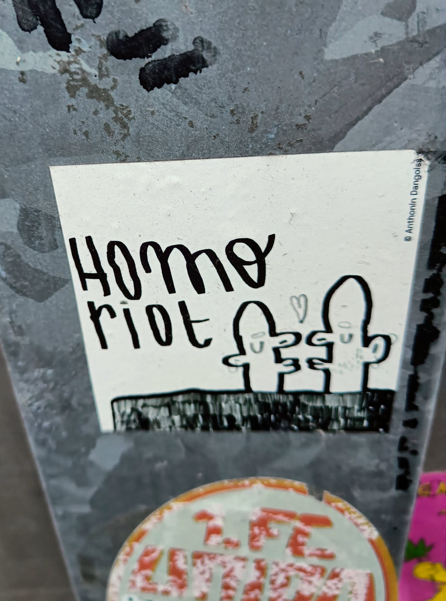 A sticker with the text "Homo Riot" and a simple drawing of figures. The sticker is white with black ink. The style is reminiscent of protest art or zine culture.