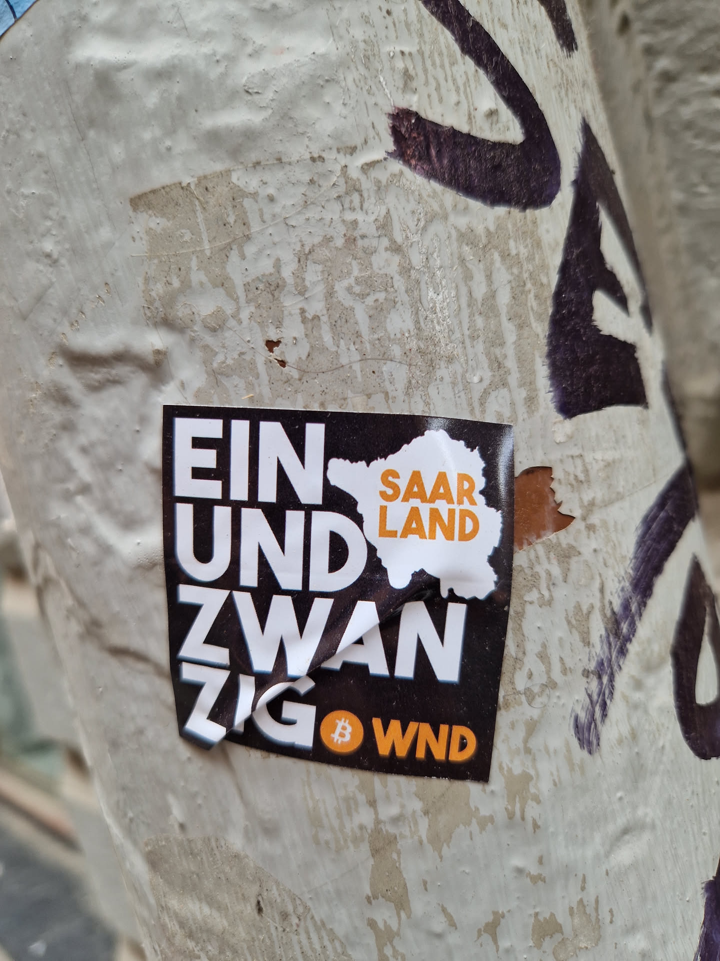 A sticker featuring the text "Ein und Zwanzig" (meaning twenty-one in German) with the Bitcoin symbol and the abbreviation "WND", likely referring to the German word "Währung" (currency).  The Saarland region map is also visible. The sticker is affixed to a weathered surface.